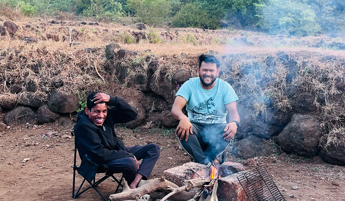 Two men sitting by a campfire, smiling and enjoying each other's company in a natural setting.
